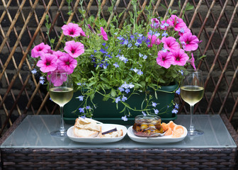 Two glasses of white wine, bread, cheese, olives, mandarin are on garden table with beautiful flowers growing in a pot. Concept: Valentine's Day invitation card.