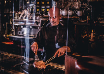 Chef cooking delicious beef steak on a kitchen, standing behind protective glass in a restaurant.