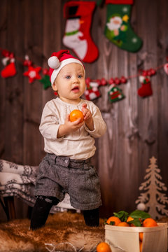 Christmas photo of the baby on a dark background of a wooden house, the baby next to the sled holds a tangerine.