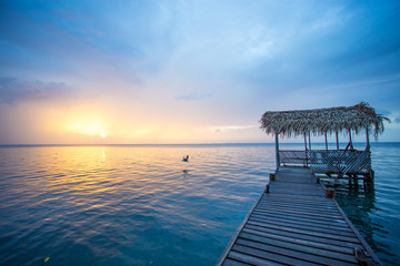 A bird is flying in the scene showing a wooden dock with a palapa roof and sunset. The water is calm and blue. - obrazy, fototapety, plakaty