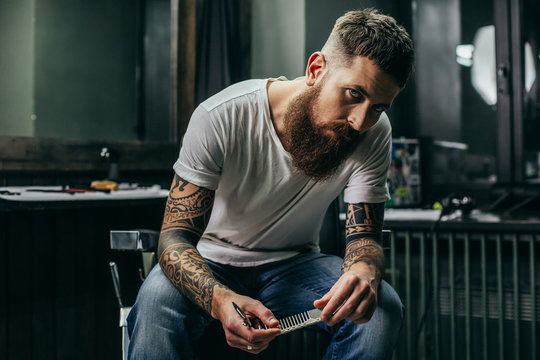 Calm bearded barber sitting and looking while holding comb
