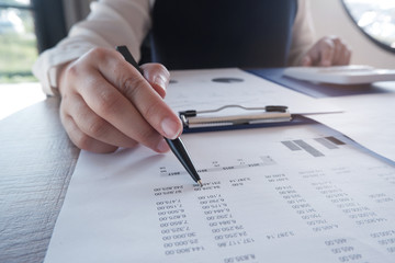 Female working in office, studying using calculator and writing something with documents and chart on table.