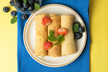 Rolled up crepes, pancakes on a plate with blueberries and strawberries. Yellow background.
