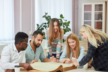 Pensive african american young man in stylish eyeglasses for vision correction, Arab man and three european young female students reading big old textbook sitting at the table against window .