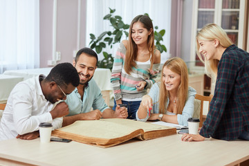 Multiethnic cheerful young people sitting at table reading big old reference book for preparing scientific report. Group of young students doing together homework at library.