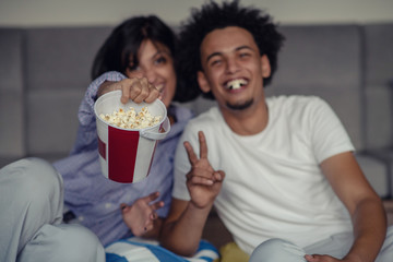 Image of smiling couple sitting on floor near sofa at home and looking aside while eating popcorn