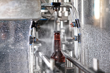 A bottle of beer on a conveyor belt under a stream of water. Only the bottle is in focus. The...