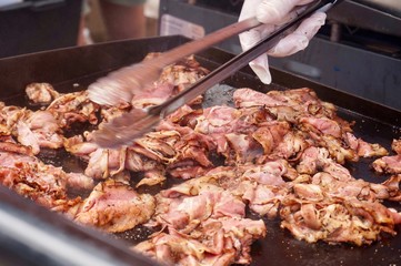 Part of gloved hand holding metal tongs and tossing meat on a flat top grill. At a food service stand at a food festival.                   