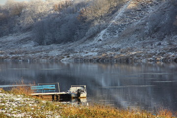 Outboard motor boat at the pier on river with a high distant shore in day at the beginning of winter on the background of the forest covered with frost - first snow, frost, beautiful water landscape