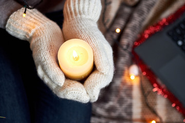Candle in hands in mittens. Christmas garland and plaid on the background. Selective focus 