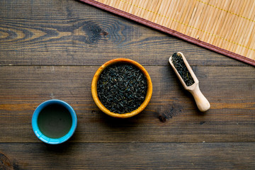 Chinese tea concept. Tea ceremony. Dry tea leaves in bowl and wooden scoop near cup of tea on rustic wooden background and bamboo table mat top view copy space