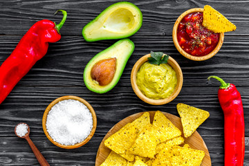 Popular mexican snack nachos. Tiangle nacho tortilla near salsa and guacamole sause, chili pepper, salt on black wooden background top view