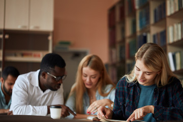 Young blonde woman writing notes with classmates studying in background. Students learning in college library.