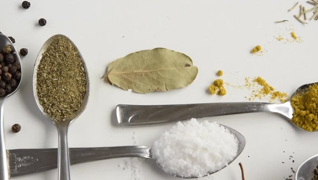 Camera moves along of spoons with various aromatic herbs and spices on white background. Different flavouring condiment choice