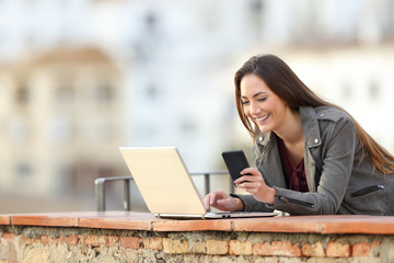 Happy woman using phone and laptop in a balcony