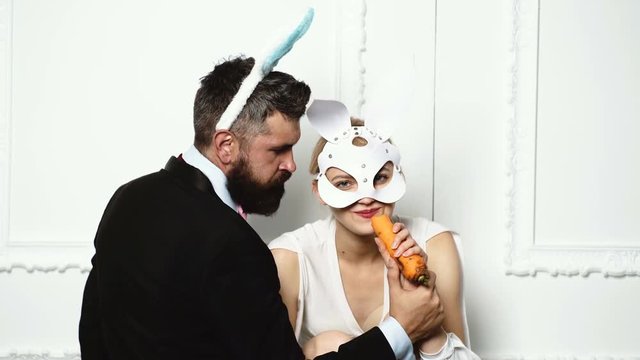 Bearded man in a suit feed woman by carrot who is dressed in a leather mask on white background. Fashion concept.