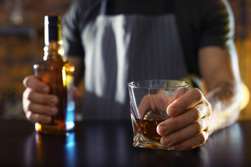 Bartender with glass and bottle of whiskey at counter in bar, closeup. Space for text