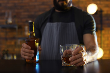 Bartender with glass and bottle of whiskey at counter in bar, closeup. Space for text