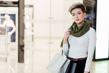 Fashionable girl with blonde haircut walking around at big well-litt shopping centre, choosing gifts for her friends and close people on Christmas day, feeling excited and happy.