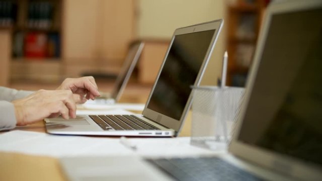 Tracking shot of unrecognizable retired woman sitting at desk and using laptop computer keyboard and touchpad