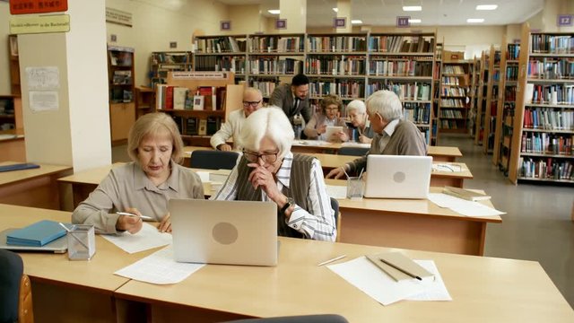 Group of active elderly people sitting at desk in library or classroom and studying while their male teacher helping two of them