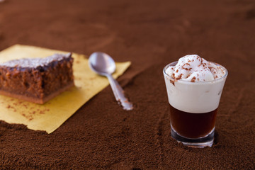 Little cup of piccolo latte macchiato on a table covered with ground coffee as a background and chocolate brownie with cocoa cream and coconut chips