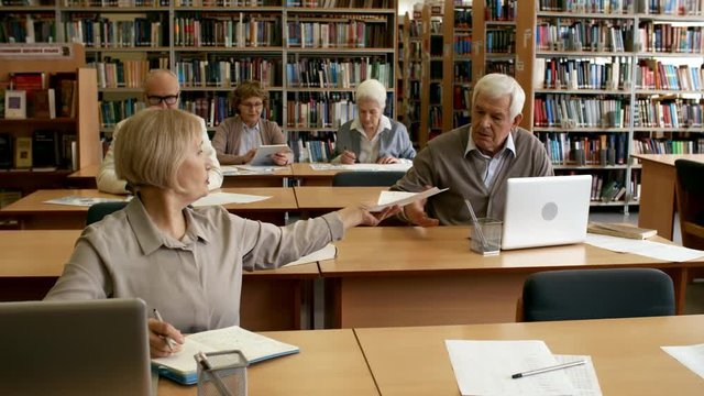 Group of elderly people studying in classroom or library, two of them passing printout with task to their group mate