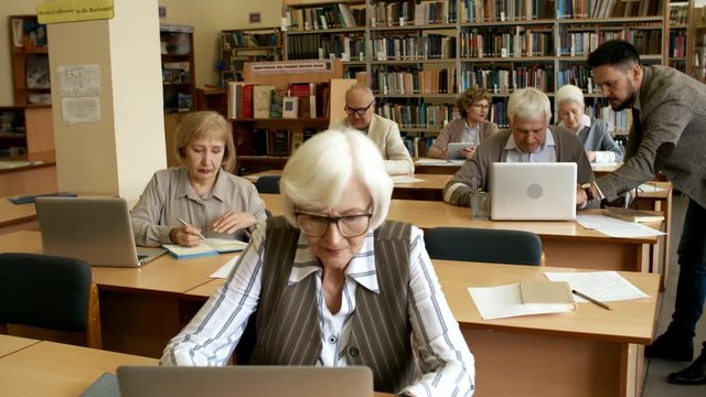 Group of elderly people sitting at desks in library or classroom and studying, middle-aged male teacher helping one of them with task