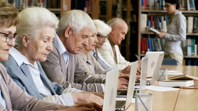 Tracking shot of group of retired people sitting next to each other in line at desks in library and using laptop computers while young female teacher speaking to them in information technology class