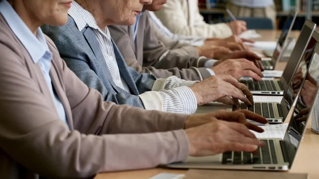 Tracking shot of unrecognizable elderly people sitting at desks and typing on laptop computers during coding class