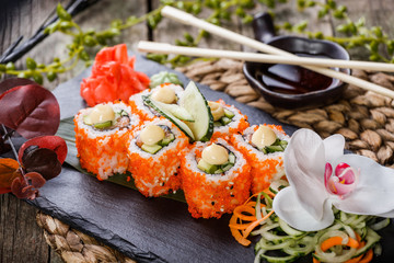 Sushi Roll - Maki Sushi made of Salmon, Red caviar, cucumber, avocado and cream cheese on black stone on bamboo mat decorated with flowers. Japanese cuisine
