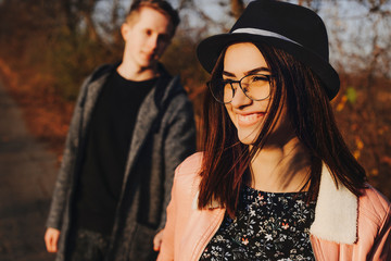 Beautiful young lady in stylish glasses and hat smiling and looking away while walking with boyfriend in autumn nature.Cheerful woman walking with boyfriend in countryside