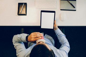 From above shot of unrecognizable male holding tablet with blank display and sipping hot drink...