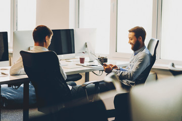 Two young men sitting on comfortable chairs and smiling while working in office together.Cheerful men working in office together