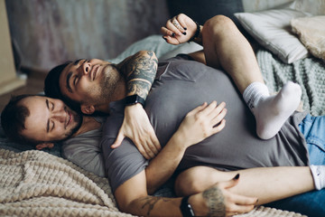 Two homosexual male partners taking care of each other. Lovely gay couple have warm cuddle, having fun in bed and smile joyfully, feeling free and natural in same-sex conduct.