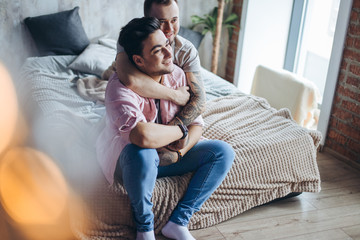 Obraz premium Happy gay couple embraced, joking and having fun in an intimate hug. Positive blue man embraces his friend, happy to be close to lover, sitting on bed at home, close up. Same-sex love relationship.