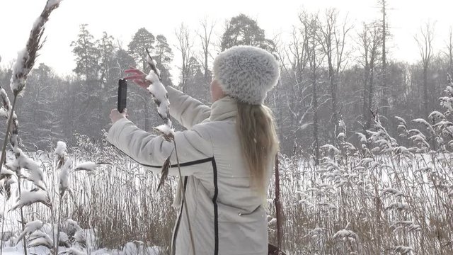 The young woman in a white jacket photographs a panorama of the coast of the winter forest lake on the smartphone,It is snowing