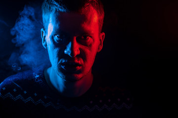 Portrait of a boy from close range who is angry and has bad emotions with a face mash and smoke around on a black isolated background. The face of the guy in the shade with color illumination.