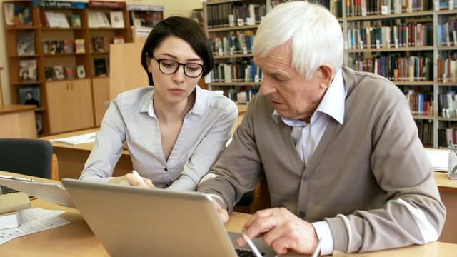 Medium shot of elderly man learning to use laptop computer in library, young female teacher helping him