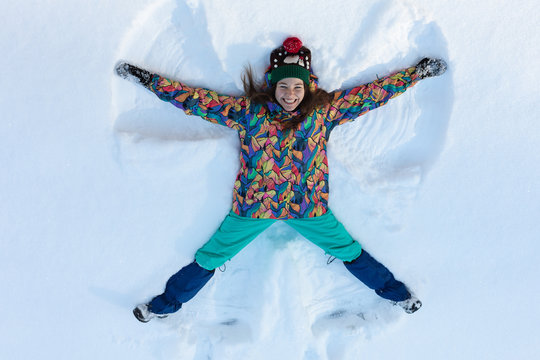 High Angle View Of Happy Girl Lying On Snow And Moving Her Arms And Legs Up And Down Creating A Snow Angel Figure. Smiling Woman Lying On Snow In Winter Holiday With Copy Space