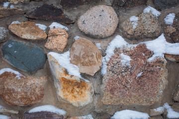 The texture is gray with a red stone wall with large stones, covered with snow. Old rough stone wall covered with snow