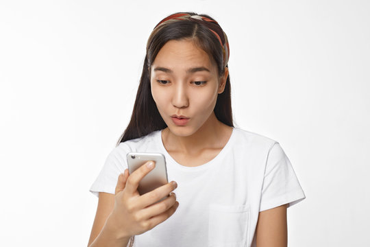 Beautiful young Chinese female with black loose hair having surprised astonished look, staring at screen of generic cell phone, watching cute video, picture or reading text message, surfing internet