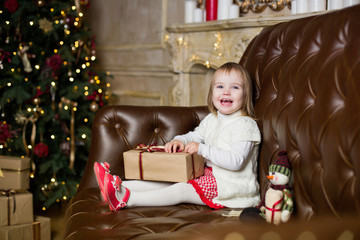 little girl in a white jacket sits on the sofa against the background of the Christmas fir, holds a gift and laughs