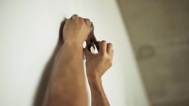Male's hands are professionally and actively driving heavy staples into white wallpapers by using staple-gun somewhere in workhouse room.