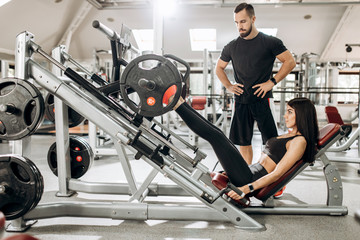 Fototapeta na wymiar Athletic girl dressed in black sport clothes is lifting weights with her legs in the gym under the supervision of a coach