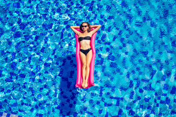 Sexy female model have a rest and sunbath on a float in the pool, top view aerial shot.woman in a black bikini swimsuit floating on an inflatable pink mattress spf and sunscreen