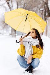 Charming dark-haired girl dressed in a yellow sweater, jeans and a white scarf is sitting on road under the yellow umbrella on a snowy  winter day