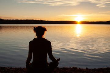 Wide shot of young woman practising yoga in nature. Meditation on the shore of the lake during sunset. Relaxation and healthy lifestyle concept.