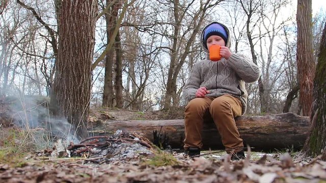 little kid in warm casual coat sitting alone on tree timber and drinking hot tea near small bonfire. Kid at family picnic in winter forest.