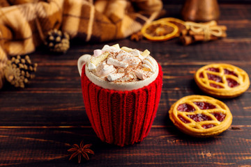 A cup of hot drink with whipped cream and powder, in a knitted cover and homemade cookies, Cezve and spices, lie on a wooden table near the checkered rug. Copy space.